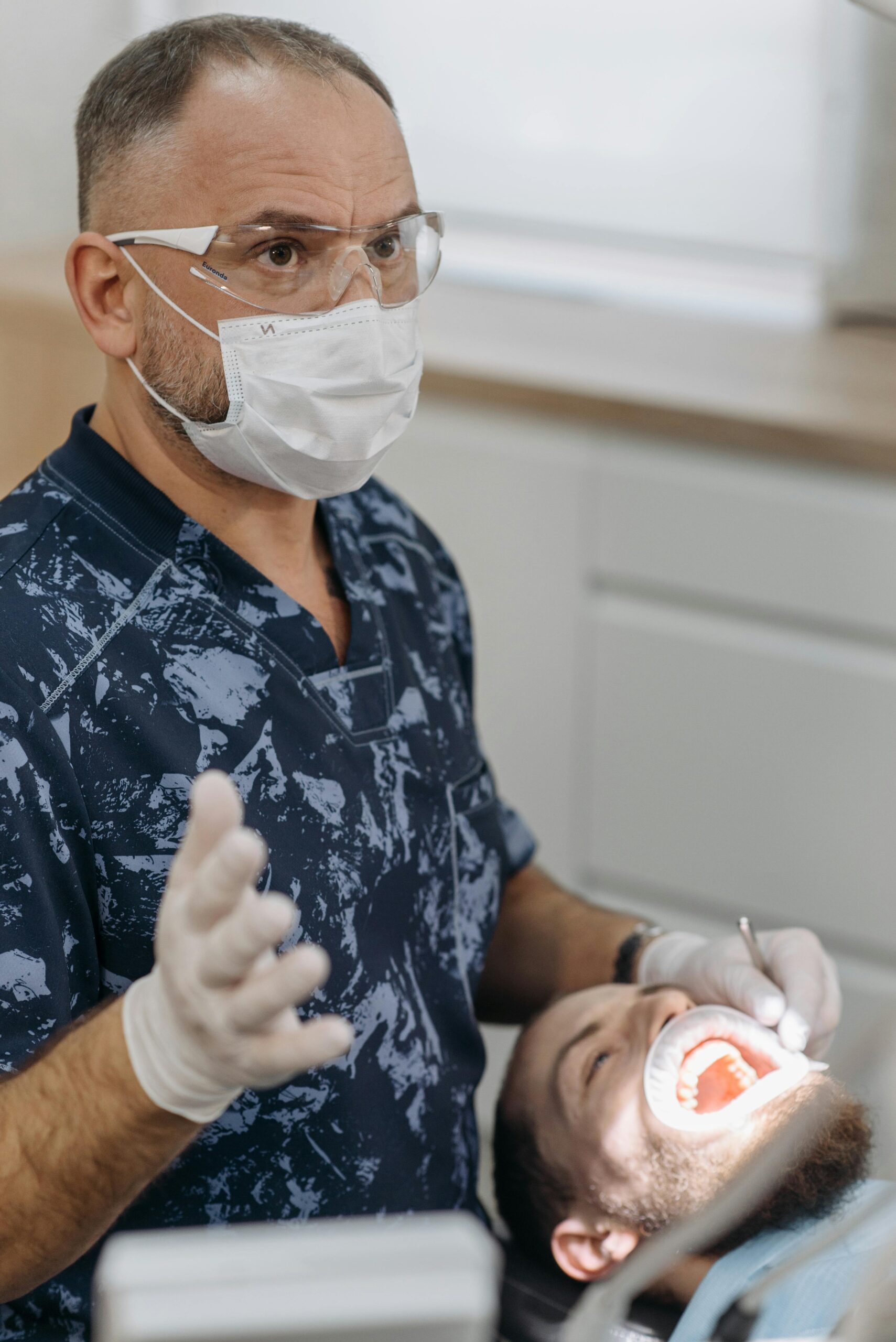 Dentist in clinic with patient undergoing dental examination, wearing protective gear.